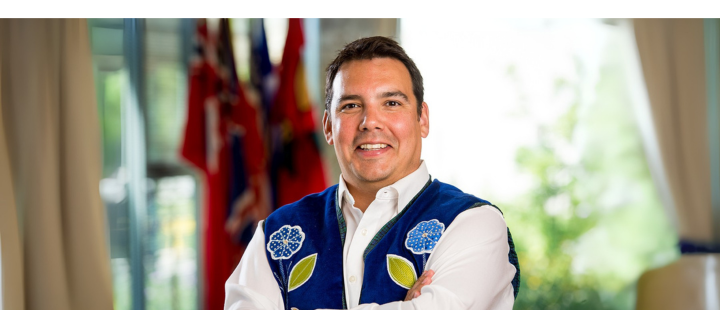 Man wearing a blue beaded vest standing with arms crossed in front of flags.