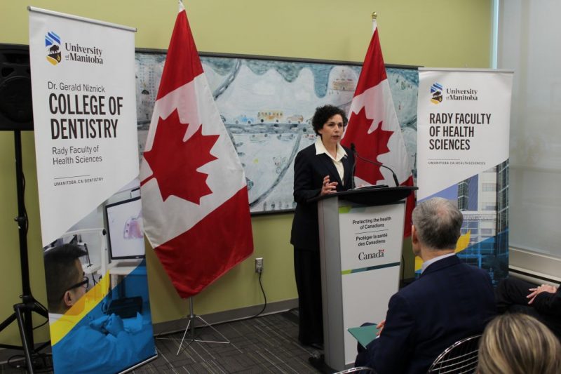 Dr. Anastasia Kelekis-Cholakis speaks at a lectern. On one side of her is a Canadian flag and a UM dental college banner. On the other side is a Canadian flag and a Rady Faculty of Health Sciences banner. 