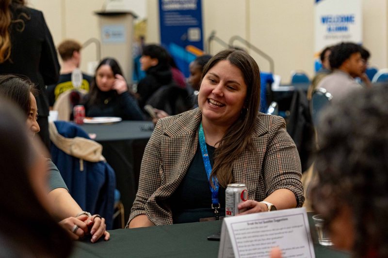 Female employer sitting at a round table with students, wearing a lanyard, smiling and chatting.
