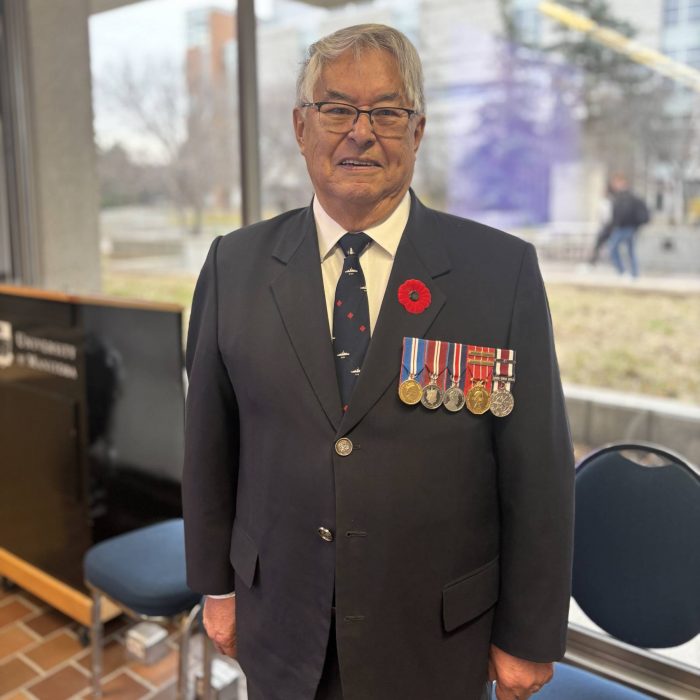 An older man wearing a dark blue suit with a white shirt and blue tie. The suit is adorned with his war metals and a red poppy. He is wearing glasses.