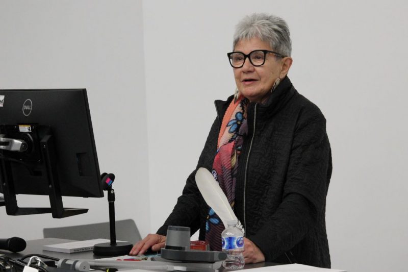 Dr. Marlyn Cook stands behind lectern with a computer monitor on it. She is holding an eagle feather. 