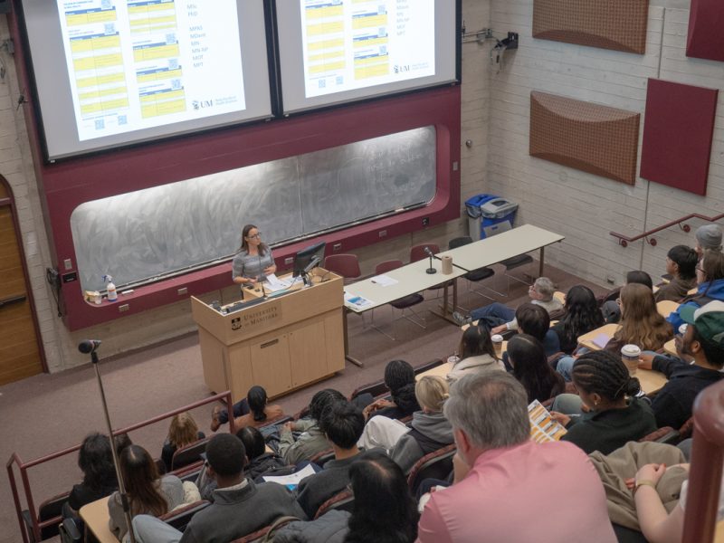 An amphitheatre style classroom is filled with students as they listen to a speaker.