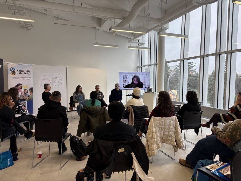 A large group of people sitting in a room watching the presenter on a TV who is presenting remotely.
