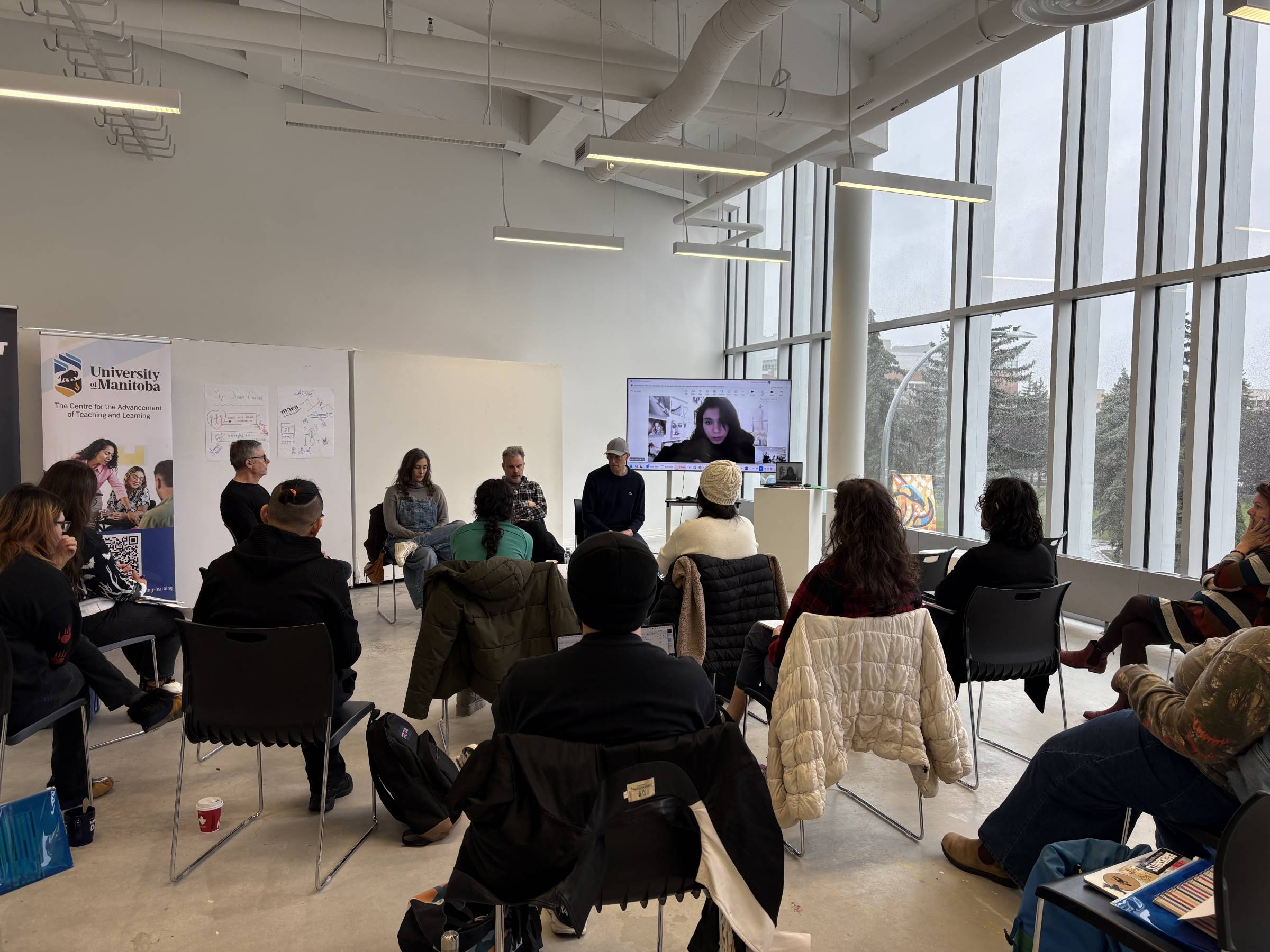 A large group of people sitting in a room watching the presenter on a TV who is presenting remotely.