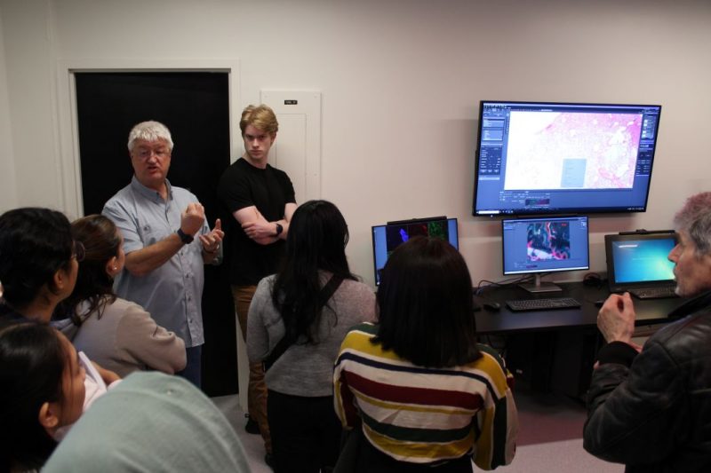 Seven people gather around Darryl Dyck. Three computer monitors and a large screen feature colourful graphics.