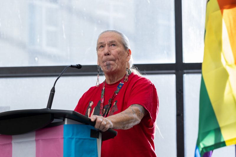 Elder Charlotte Nolin speaks at a podium, which has a trans flag on front.