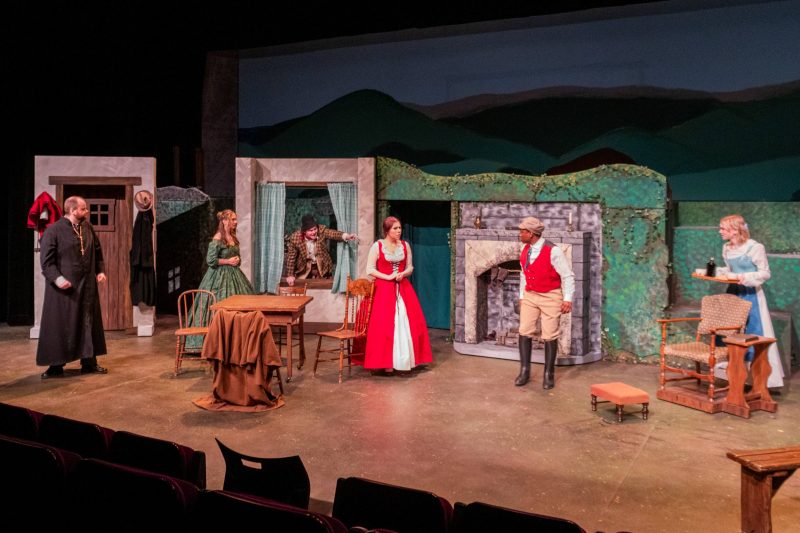 Six student actors performing on stage wearing period costumes from the 19th century.
