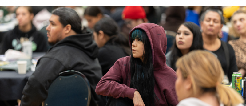 A large group of people sit closely together at round tables during an event. A young woman in a maroon hoodie sits in the foreground with her knee pulled up, looking attentively toward the front.
