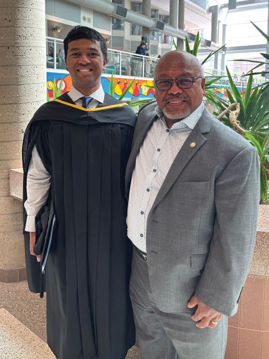 Nick and Tom pose for a photo. Nick is wearing a graduation gown.
