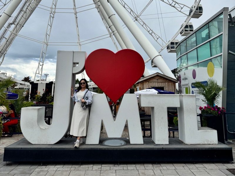 Gloria Lee standing beside a J’ ❤ MTL sign with a ferris wheel in the background.