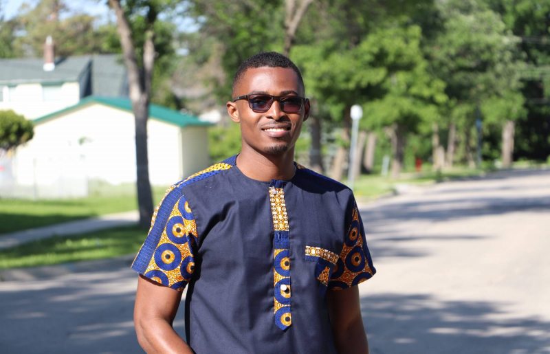 Young Black man outdoors with green trees in background.