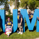 Five people smiling around big blue "UM" letter sign outdoors