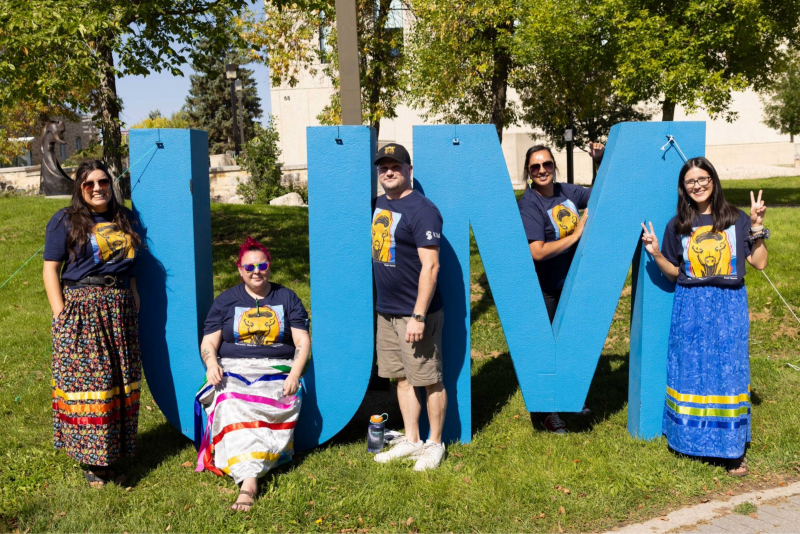 Five people smiling around big blue "UM" letter sign outdoors