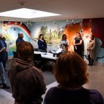 Students and community members gather in the Indigenous Student Space for opening remarks, surrounded by a mural.