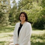 Tréchelle Bunn in a white blazer standing in an open green space surrounded by trees, sunshine and blue sky.