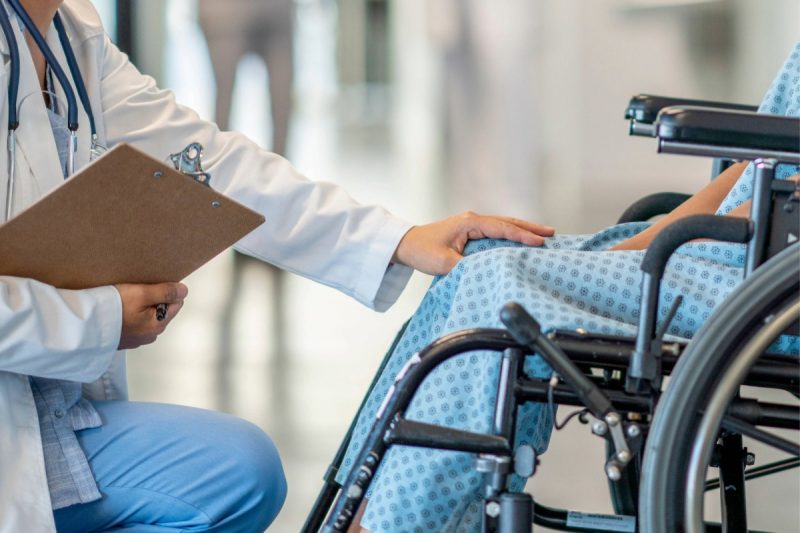 A patient sits in a wheelchair and is helped by a doctor who approaches patient in an empathetic way.