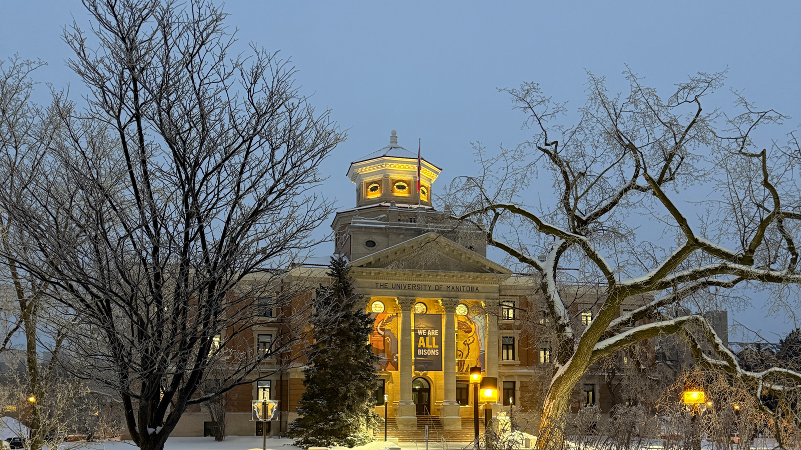 Snow covered UM Admin building from the patio of UMSU University Centre, with snow covered trees in the foreground on the mild and cloudy morning of January 5, 2026 - Zachary LeClerc