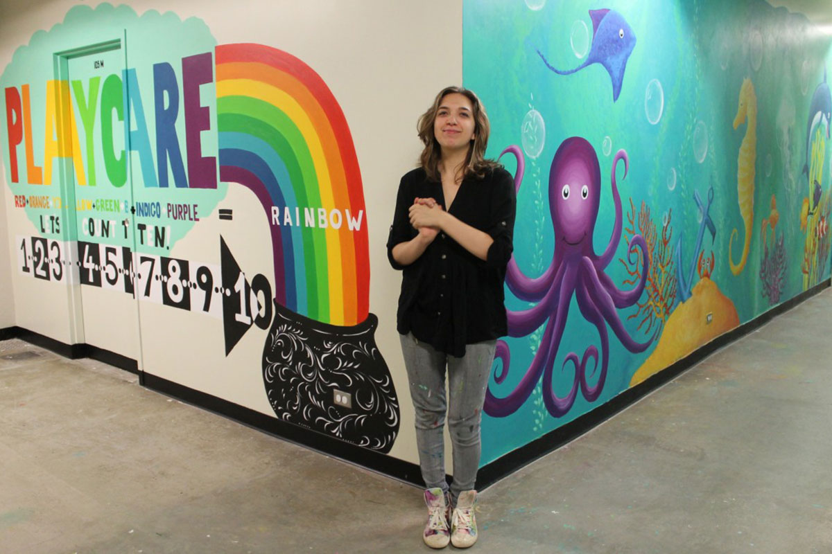 Mural Artist Anya Nikoulina stands next to the mural that welcomed children and parents to the PlayCare Centre in University College.