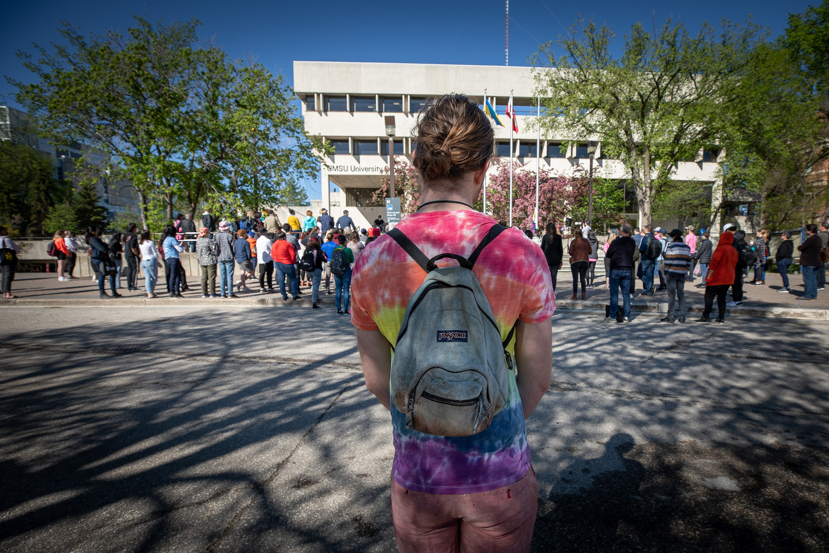 Person with rainbow tie-dye t-shirt watches the Pride flag raising wtih a group of people.