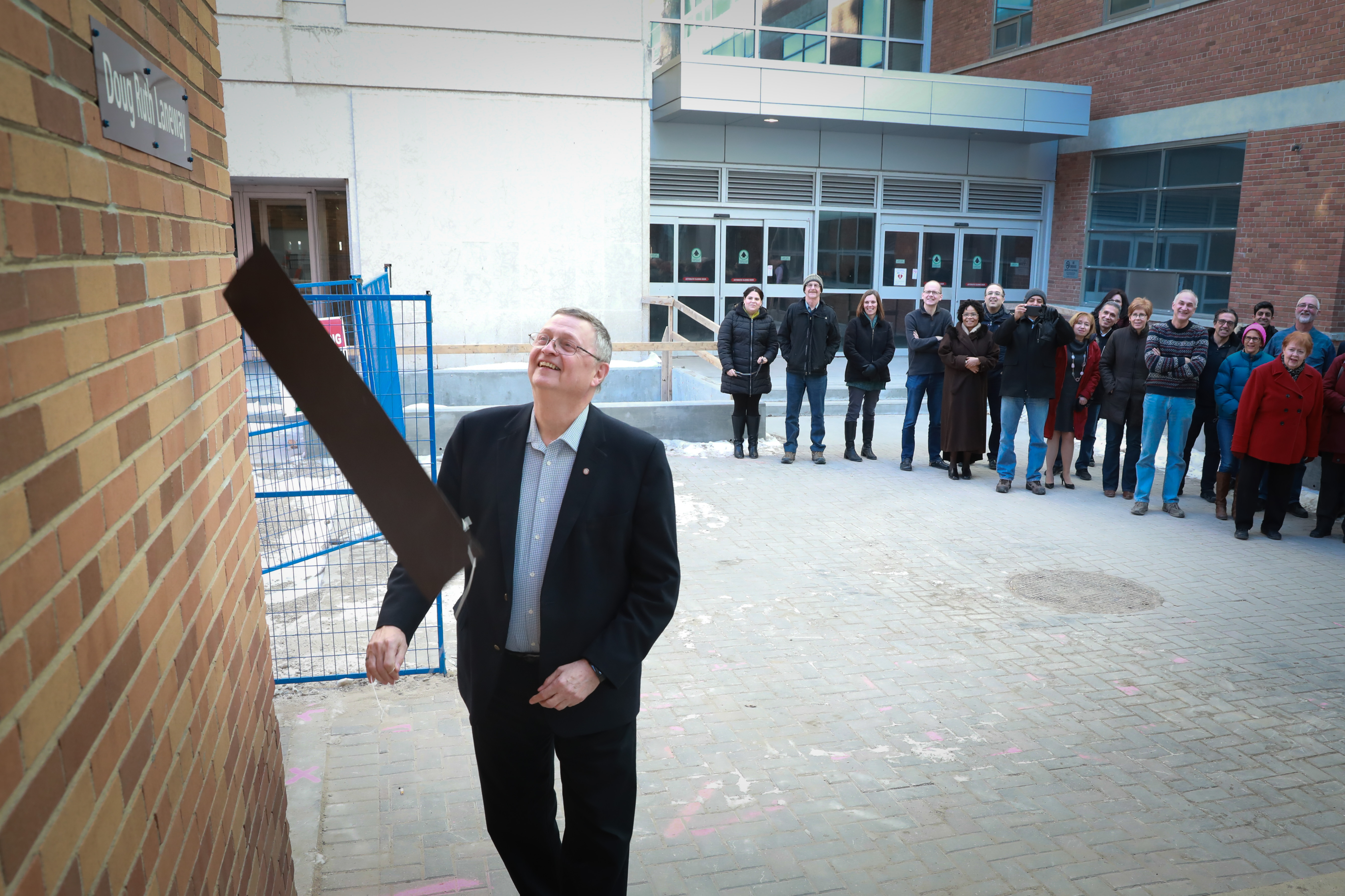 Alt: Smiling, Doug Ruth pulls off a magnetic strip from a brown sign with white letters,  reveling "Doug Ruth Laneway"
